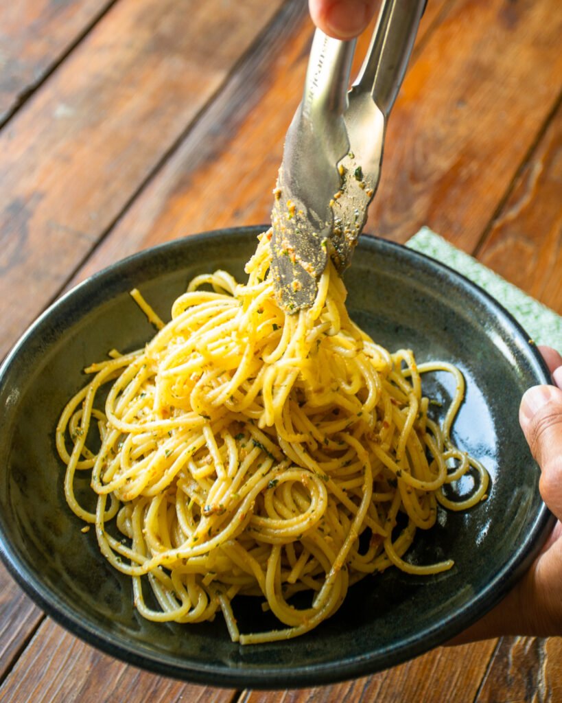Tongs placing sun-dried tomato pesto spaghetti into a black bowl.