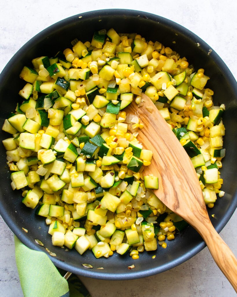 Zucchini, corn and onions in a skillet with a wooden spoon.