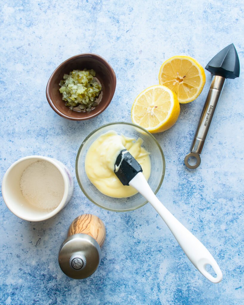 All the ingredients for tartar sauce on a blue countertop with a spatula and a reamer.