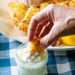 A hand dipping a fish stick into a jar of tartar sauce with a plate of fish sticks in the background.