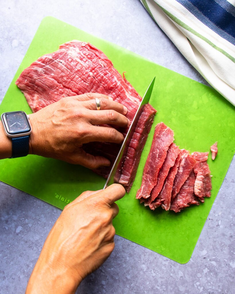Hands slicing flank steak into strips on a green cutting mat.