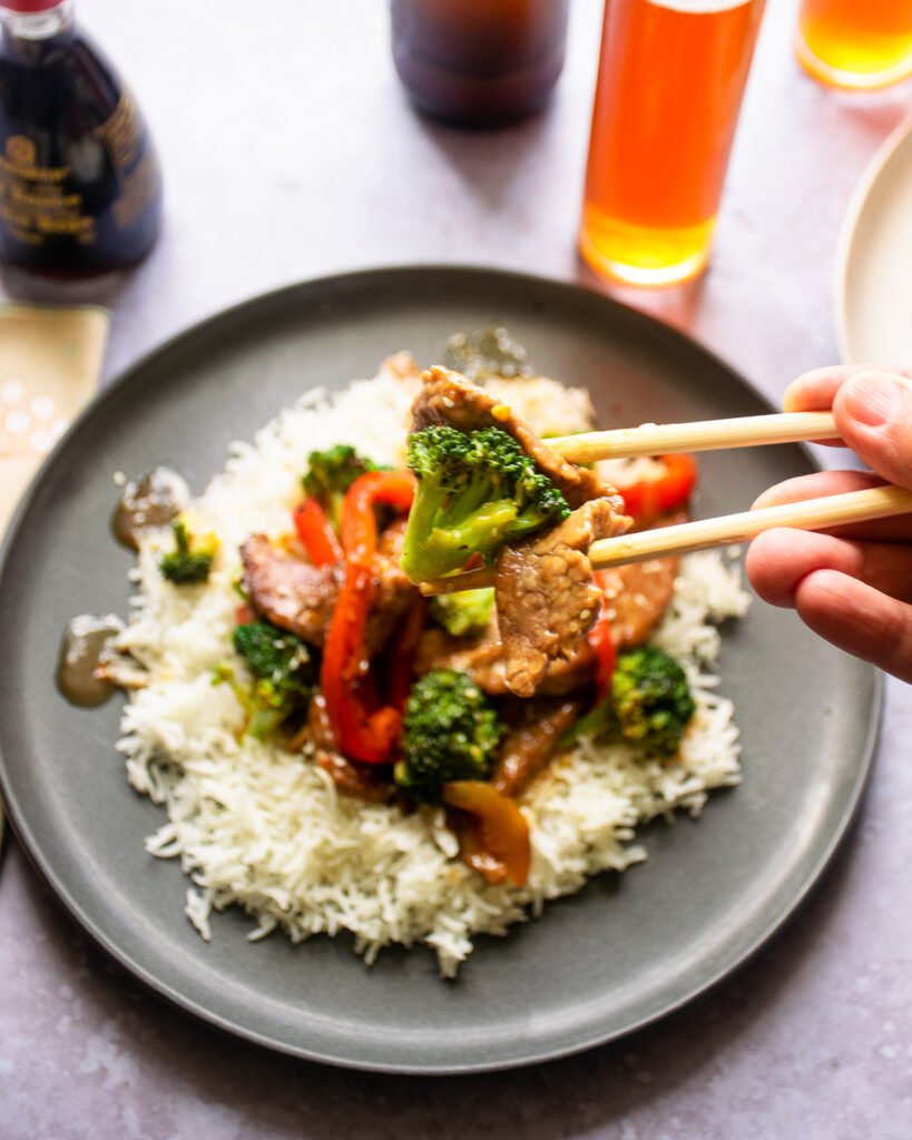 Hands using chop sticks to pick up a piece of broccoli from a plate of stir fry on rice.