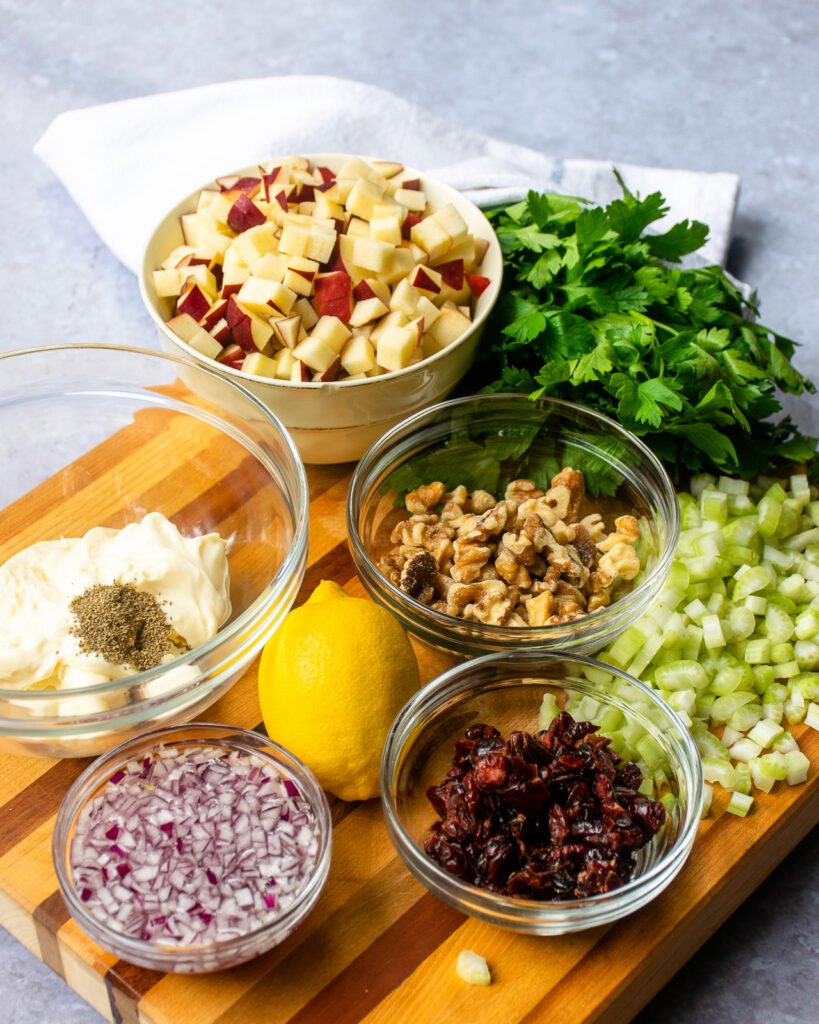 Ingredients on a wooden cutting board - red onions, cranberries, celery, walnuts, a lemon, parsley, chopped apple and a bowl of mayonnaise.