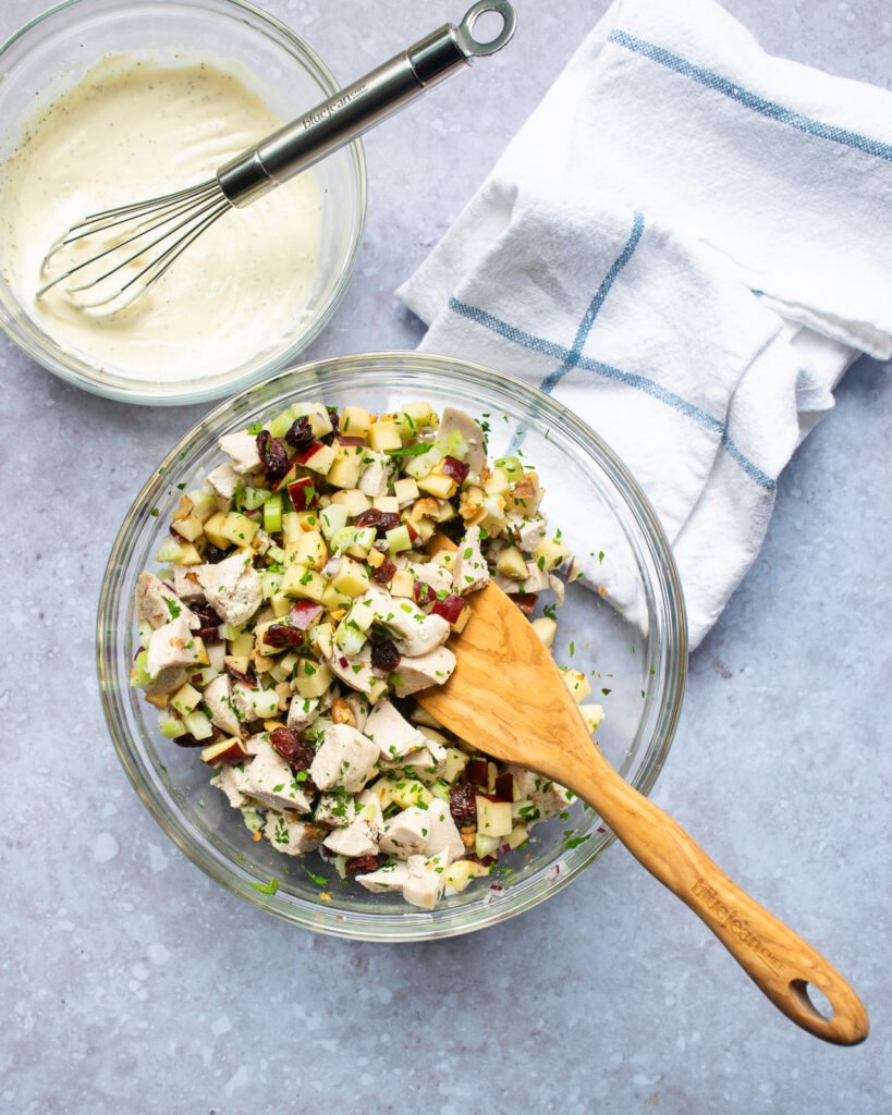 A bowl of chicken salad ingredients with a wooden spoon sticking out of it next to a second bowl with a mayonnaise dressing and whisk.
