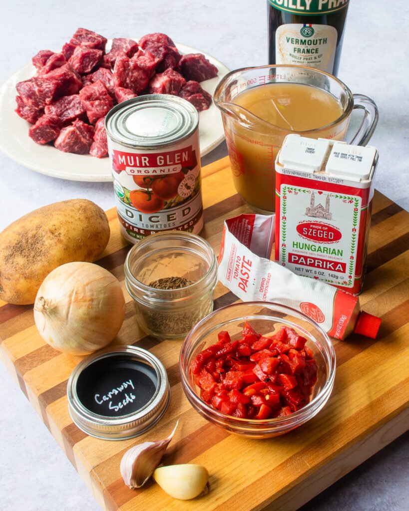 Ingredients on a cutting board - paprika, carraway seed, onion, potato, roasted red peppers, canned tomatoes, tomato paste, stock and stew beef.