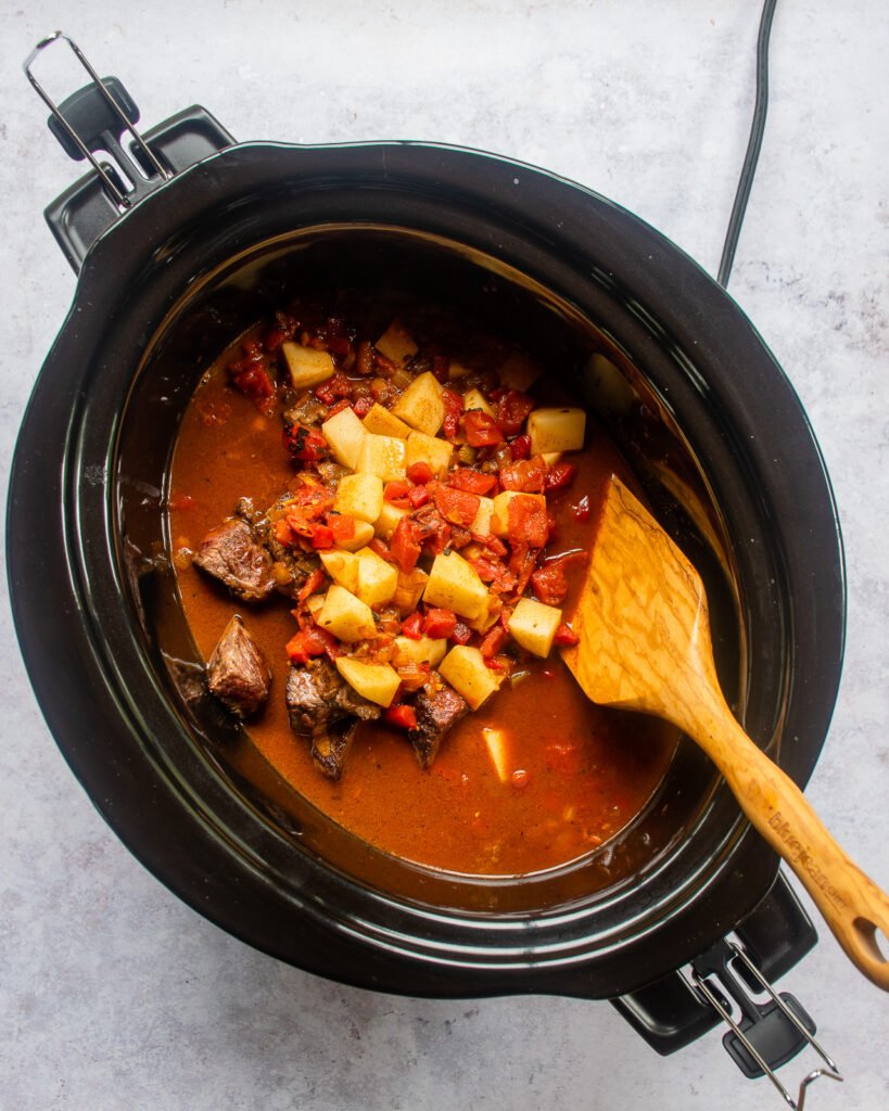 Ingredients for Hungarian Goulash in a slow cooker with a wooden spoon sticking out of it.