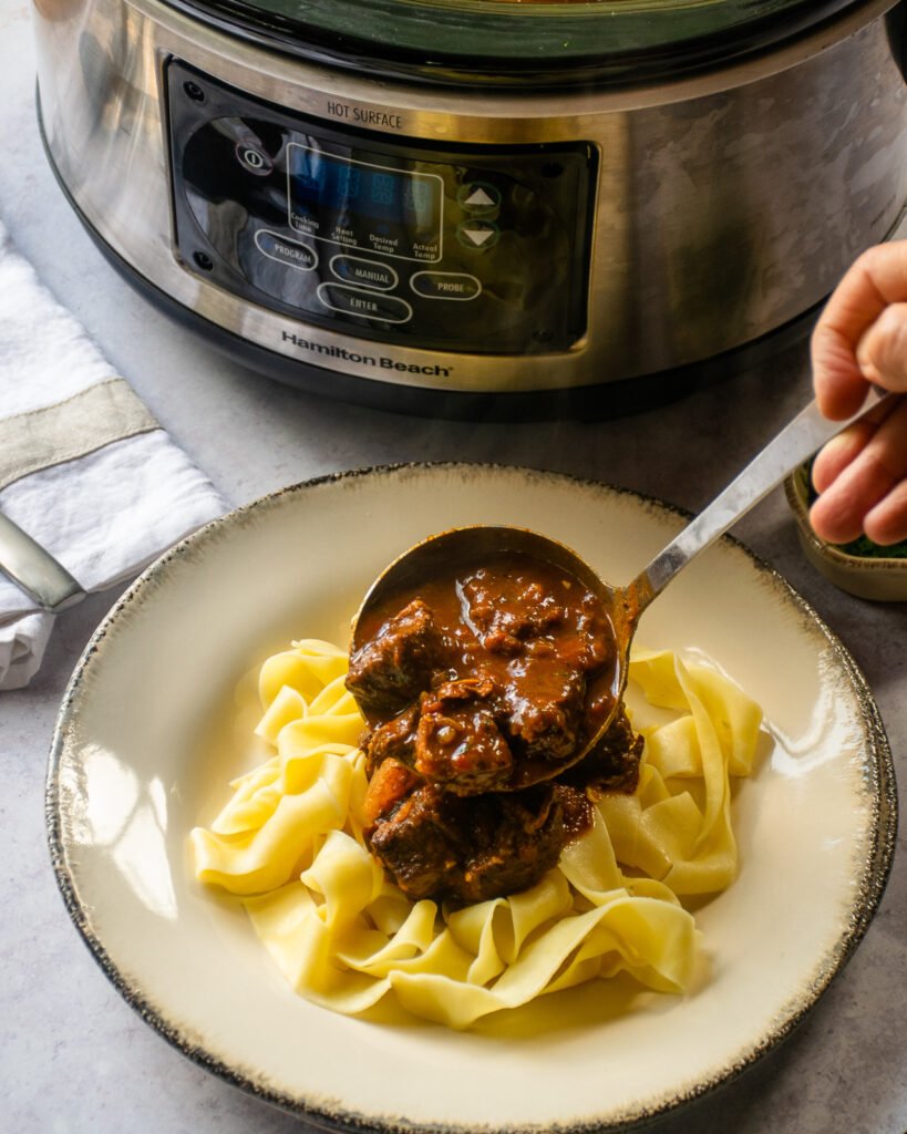 A hand ladling Hungarian Goulash over egg noodles with a slow cooker in the background.