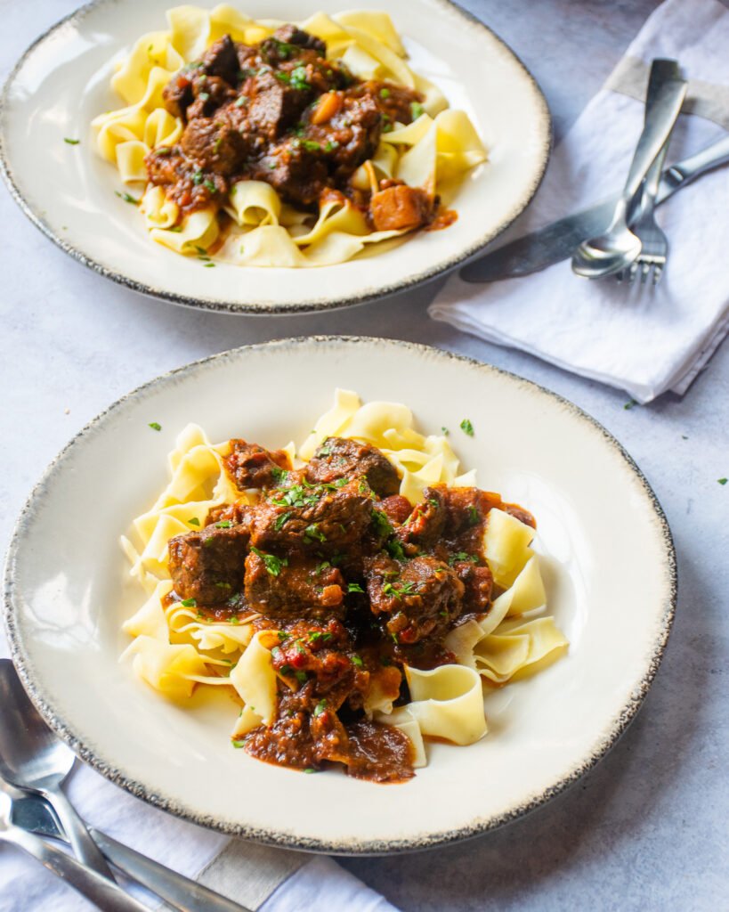 Two plates of Hungarian Goulash on egg noodles on a tabletop with cutlery..