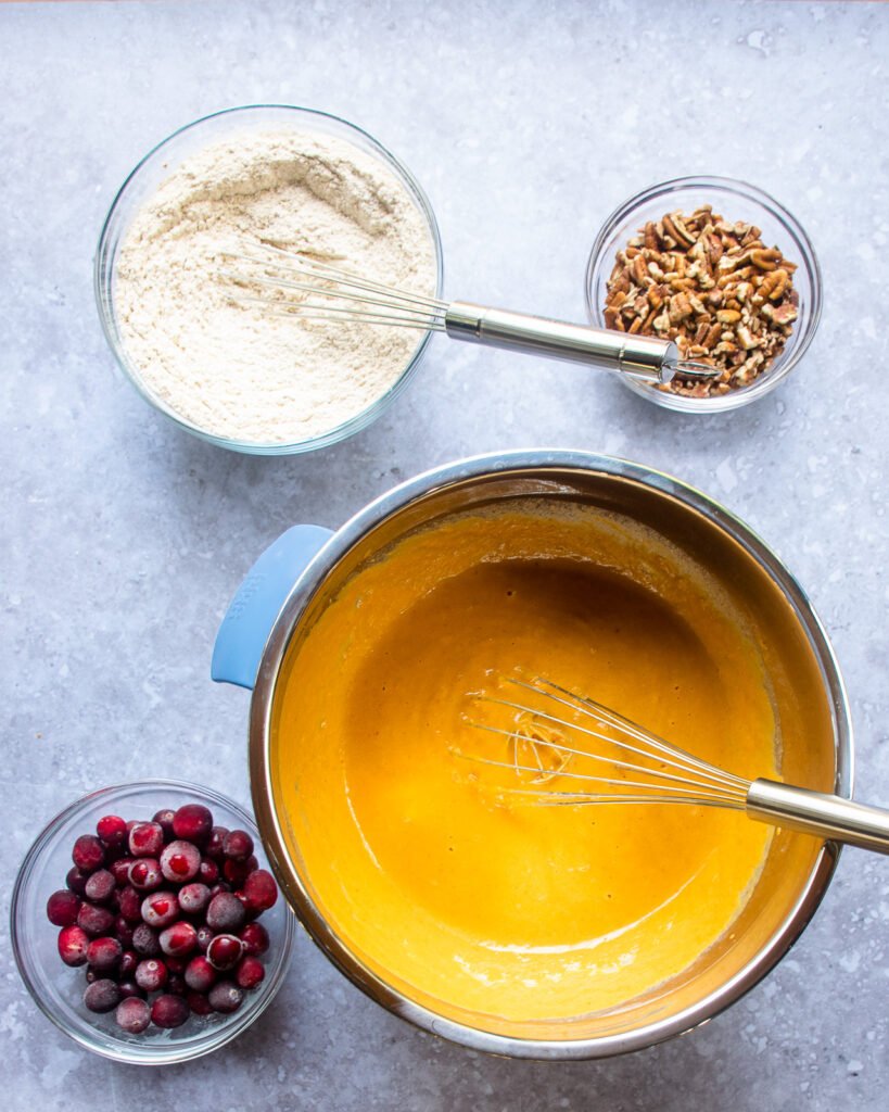 Looking down on four bowls - one with cranberries, one with pecans, one with flour and dry ingredients and a fourth with a wet orange batter.