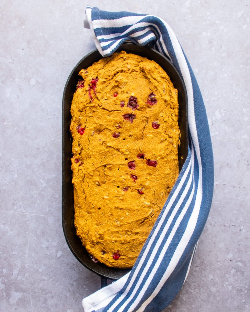 Pumpkin cranberry cornbread in an oval cast iron pan with a blue and white striped towel.