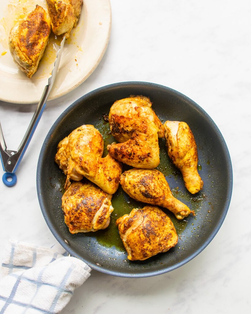 Chicken pieces browning in a skillet with a plate of browned chicken next to it with a pair of tongs.