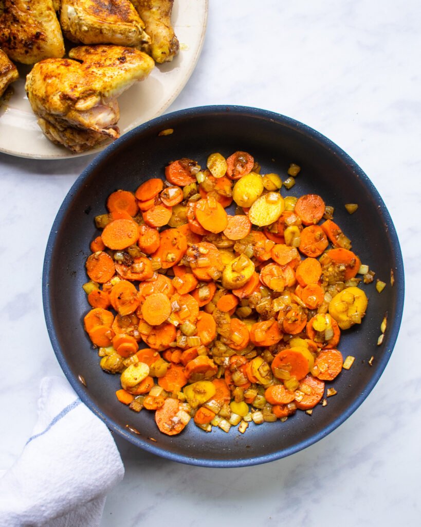 Looking down on a skillet of vegetables next to a plate of browned chicken.