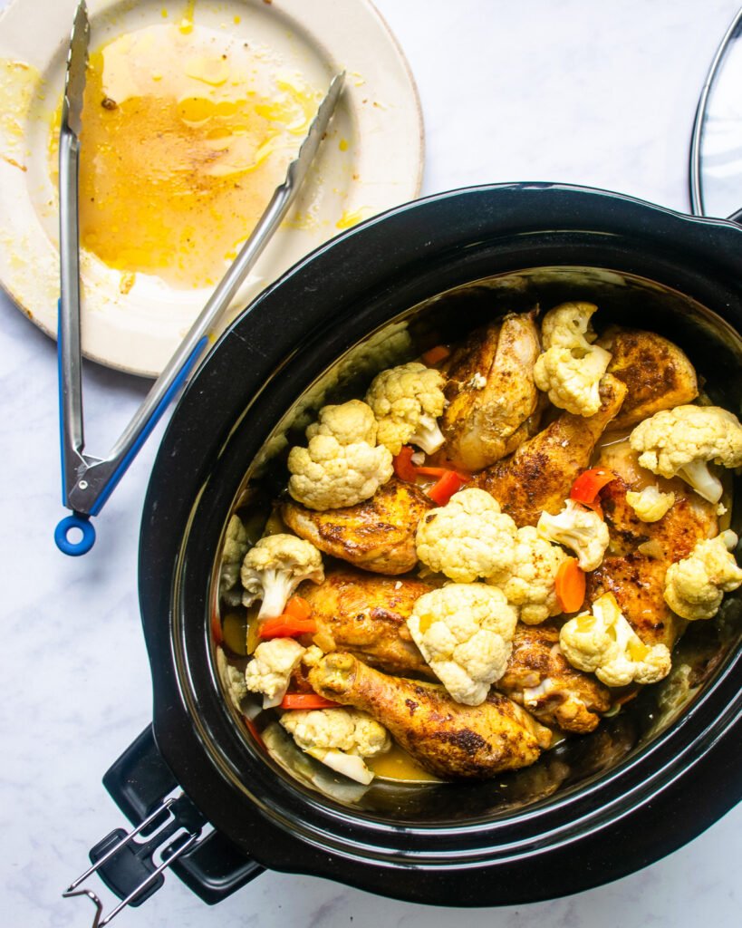 Slow cooker curried chicken stew in a slow cooker before being cooked. An empty plate and tongs near by.