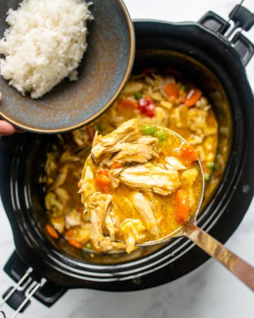A ladle of curried chicken stew coming out of a slow cooker and towards a bowl of rice.