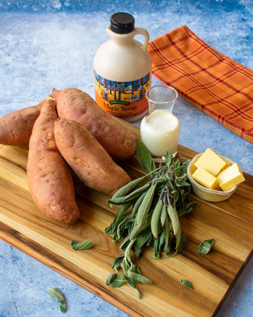 Ingredients on a cutting board - sweet potatoes, sage, butter, cream and maple syrup with an orange kitchen towel in the background.