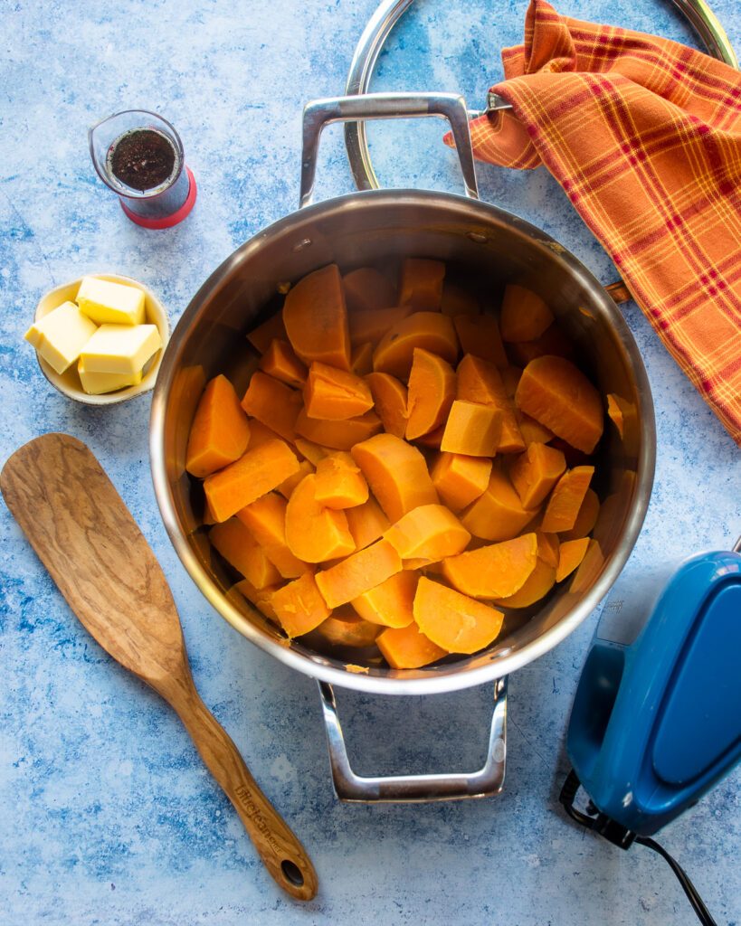 Looking down into a stainless steel stockpot with cooked sweet potato chunks inside, butter, maple syrup, a wooden spoon and a hand mixer next to it.