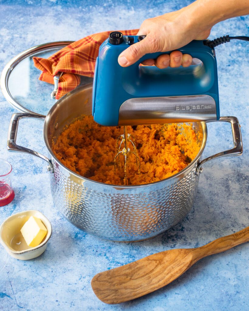 A hand using a hand mixer to mash sweet potatoes in a stainless steel stockpot.