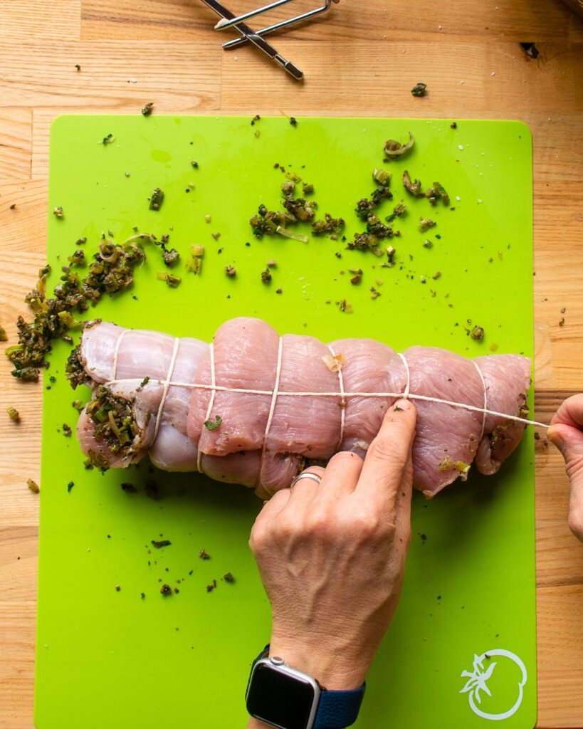 Two hands showing how to tie up a roast turkey breast., showing how to thread the end of the string over and under the perimeter twine loops.
