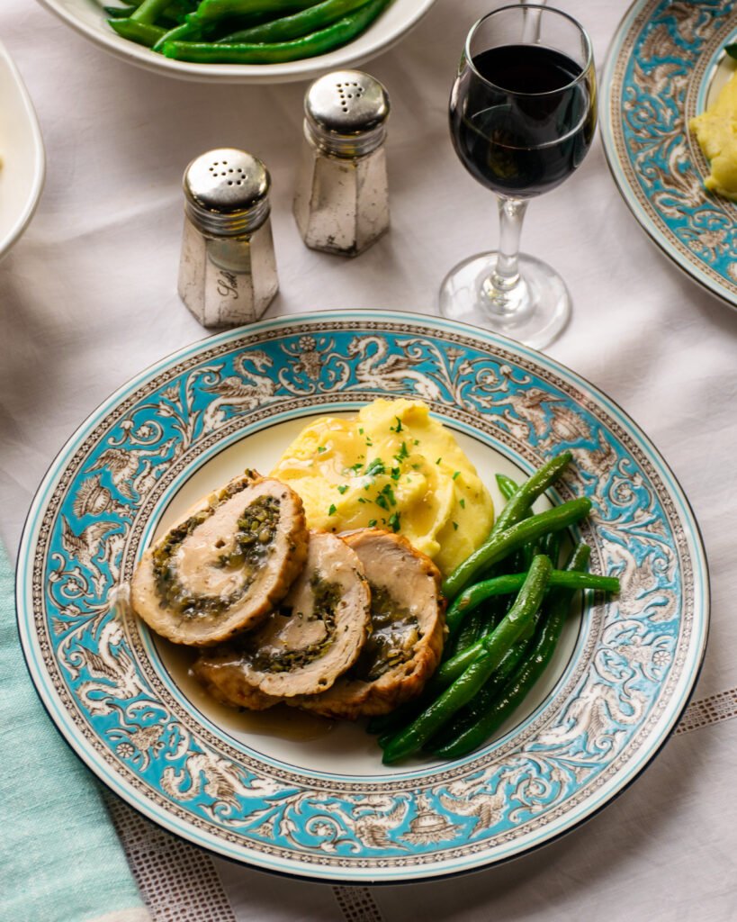 A china dinner plate of turkey roulade, gravy, green beans and mashed potato on a white table cloth with a second place setting, wine and salt and pepper shakers.