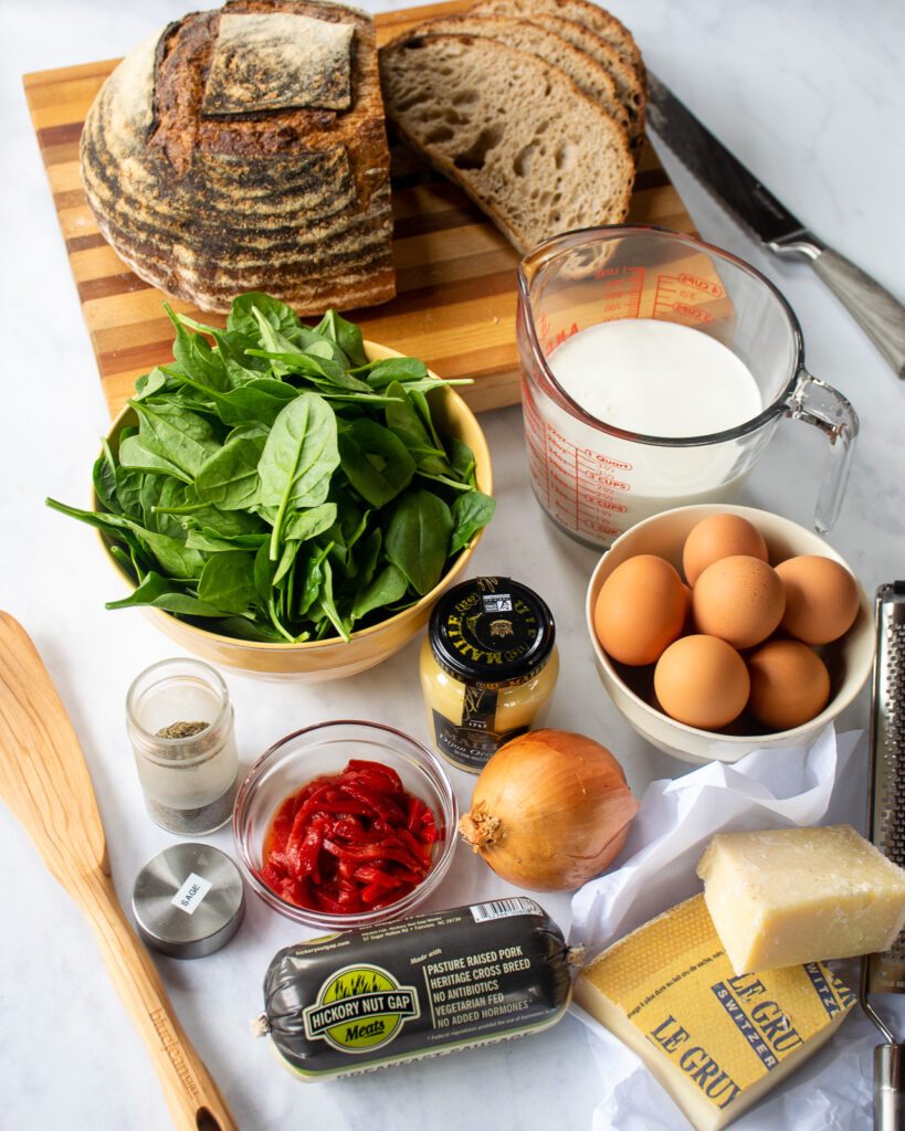 Ingredients for breakfast strata - sausage, cheeses, red peppers, dried sage, mustard, onion, eggs, spinach, half-and-half and a loaf of sourdough bread.
