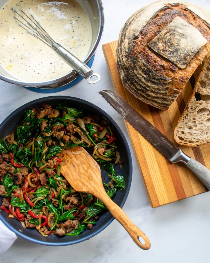 Looking down on a loaf of sourdough bread with bread knife, a skillet with sausage and spinach filling with a wooden spoon and a bowl of egg custard with a whisk.