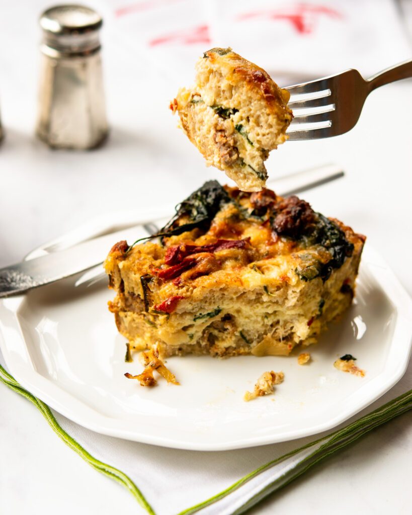 A fork lifting out a bite of breakfast strata from a portion on a white plate.