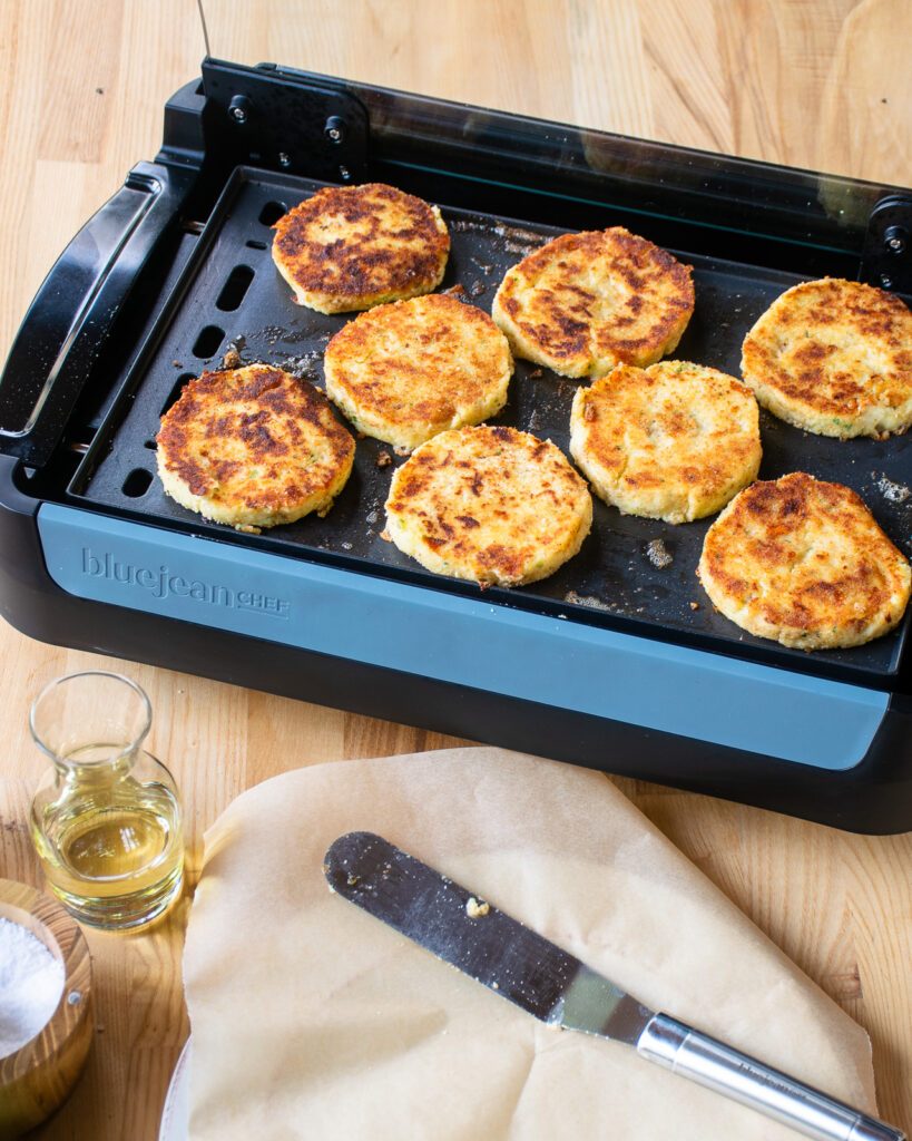 Mashed potato cakes cooking on an electric griddle with a parchment lined plate and metal spatula in front of it.