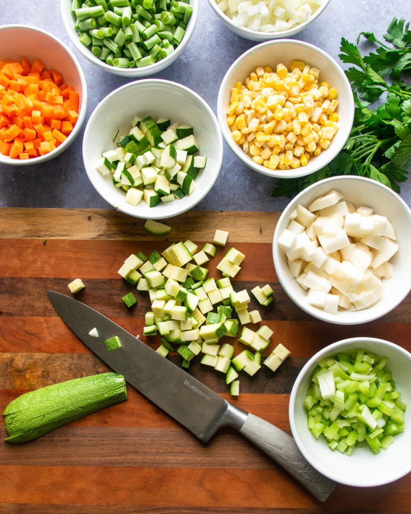 A knife cutting zucchini into cubes with small white bowls of other ingredients nearby.