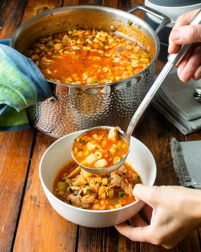 Hands ladling chicken vegetable soup from a hammered stainless steel stockpot to a white bowl.