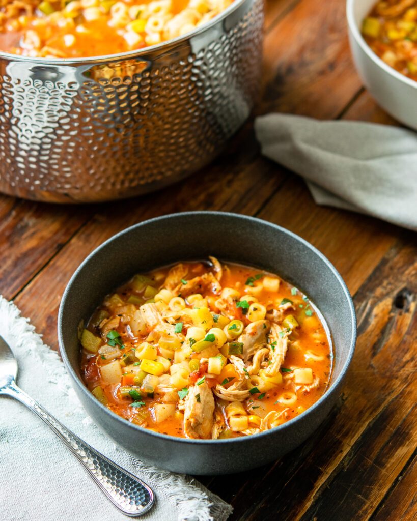 A dark gray bowl of chicken vegetable soup with a stockpot and another bowl of soup in the background.