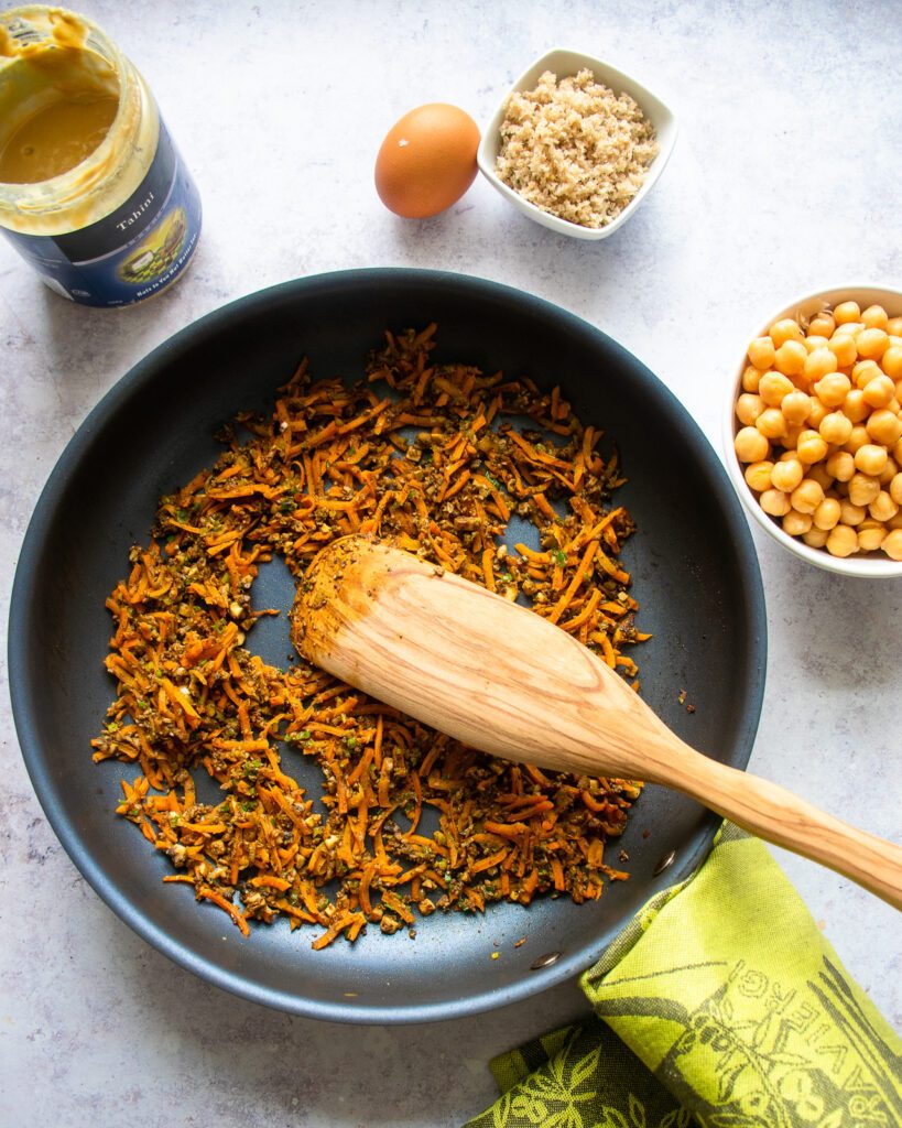 Vegetables cooking in a large skillet with a wooden spoon resting in it, and tahini, chickpeas, breadcrumbs and an egg on the counter next to it.