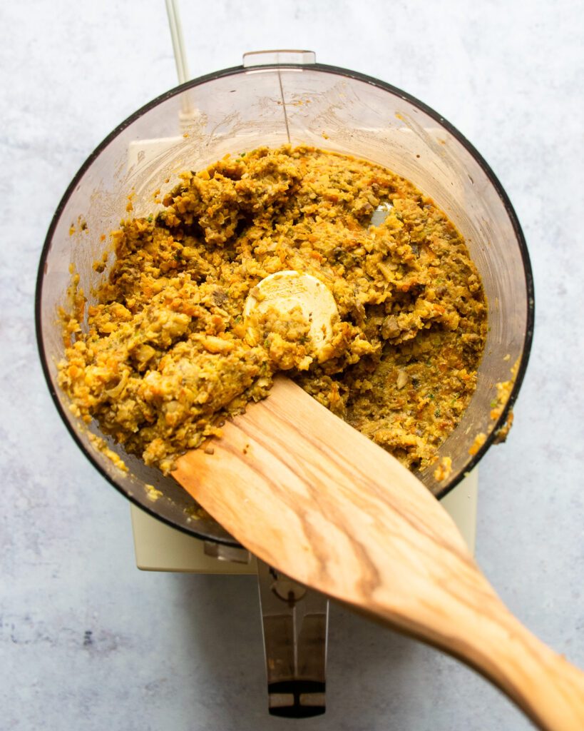 Chickpea burger mixture in a food processor with a wooden spoon scooping some out.