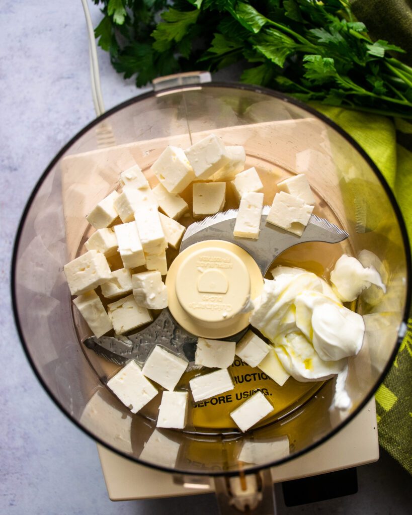 Looking down into a food processor with feta cheese, yogurt, olive oil inside and fresh parsley on the counter next to it.