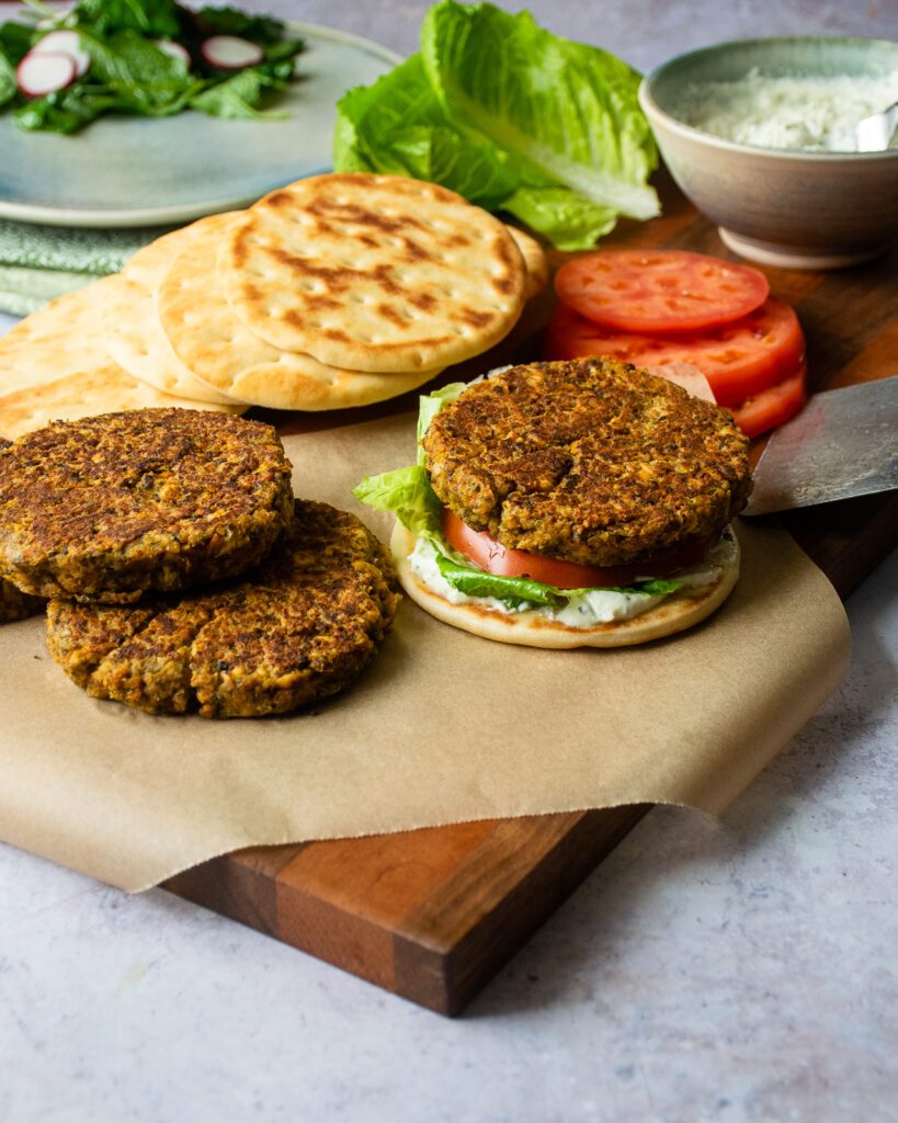 Chickpea burgers being assembled on a wooden board, with naan minis, tomato slices, lettuce and feta spread in the background.
