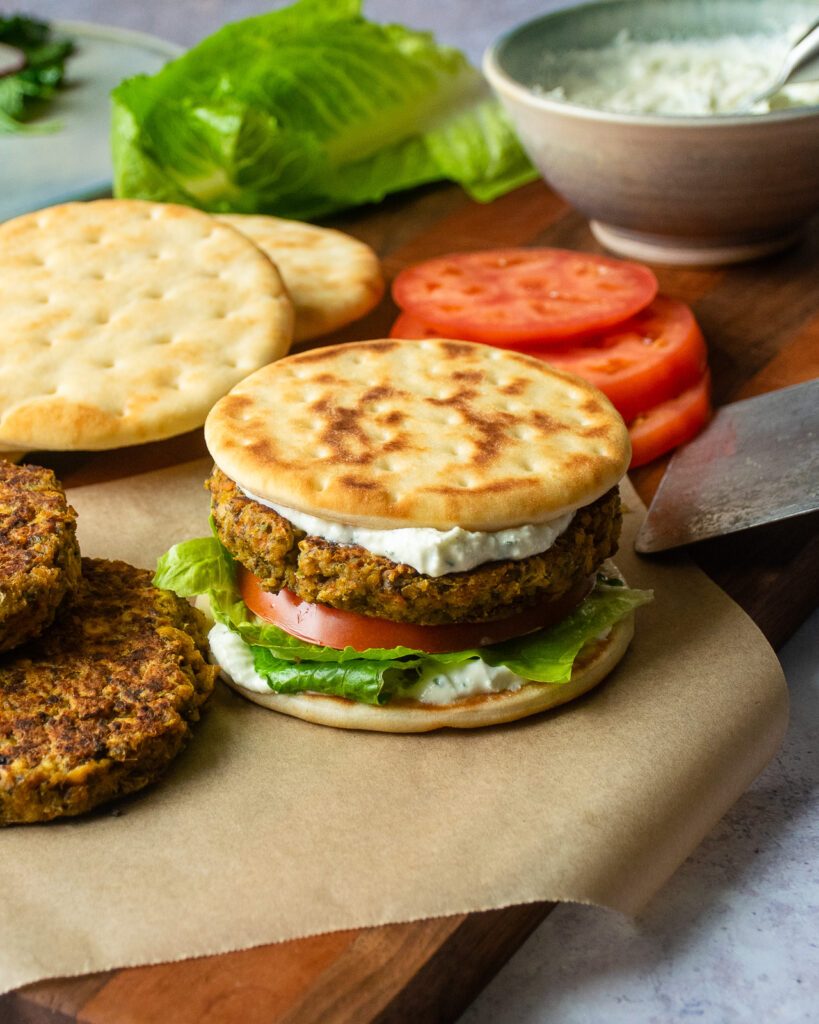 Chickpea burger on parchment paper with lettuce, tomato, feta spread and more burger patties nearby.
