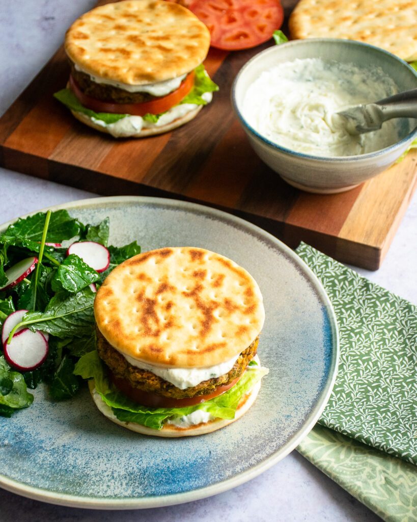 A chickpea burger on a blue plate with a side salad, and more chickpea burgers and whipped feta spread in the background.