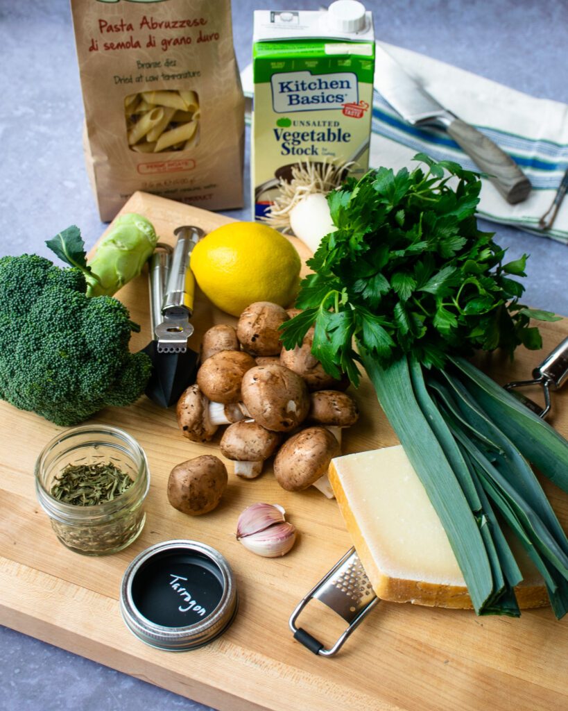 Ingredients on a cutting board - mushrooms, parmesan cheese, a leek, parsley, broccoli, lemon, dried tarragon, pasta and vegetable stock.