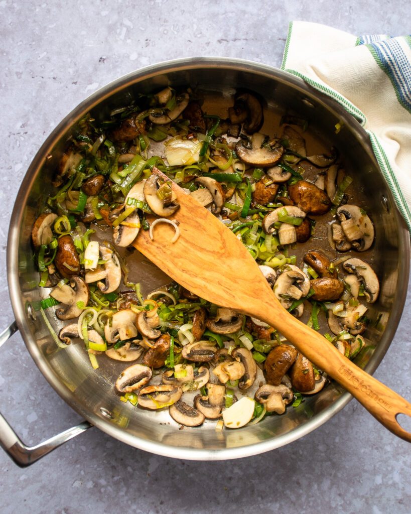 Mushrooms and leeks sautéing in a large sauteuse with a wooden spoon.
