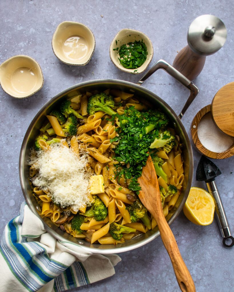 Pasta with mushrooms, broccoli and leeks in a large sauteuse with parmesan cheese and parsley on top, before being stirred.