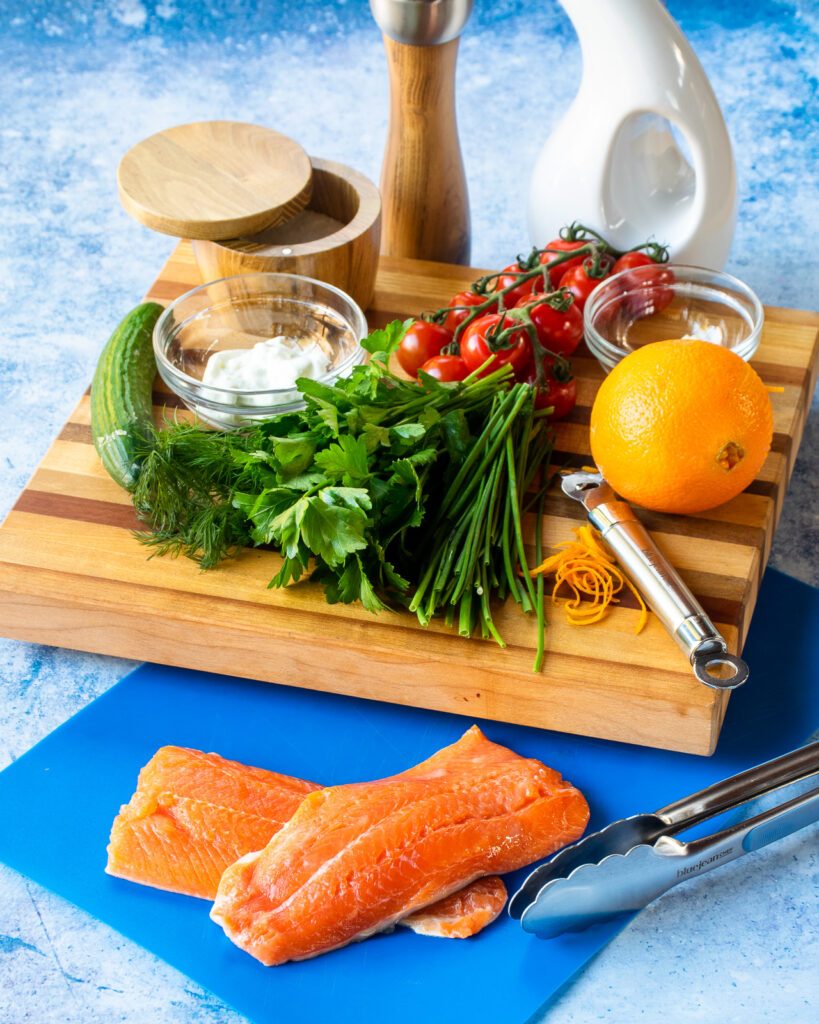 Ingredients on a blue counter - arctic char fillets, herbs, an orange, tomatoes, salt, pepper and olive oil.
