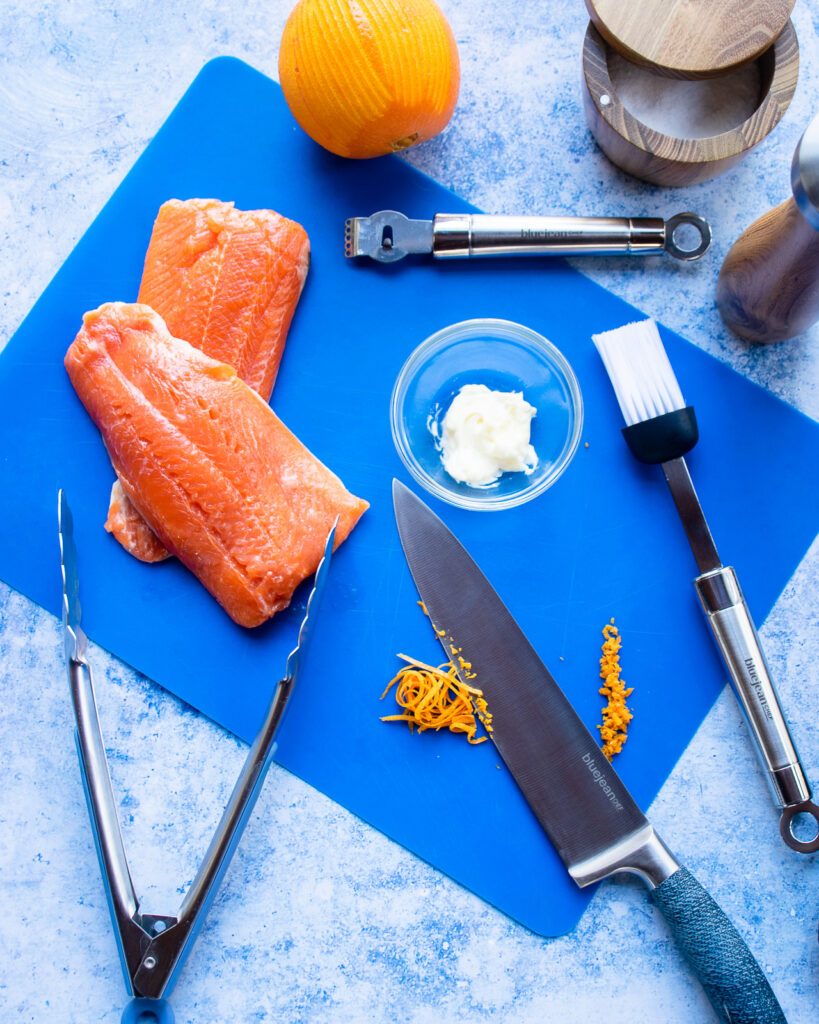 Looking down on a blue cutting mat with arctic char fillets, a bowl of mayo, an orange and some orange zest.