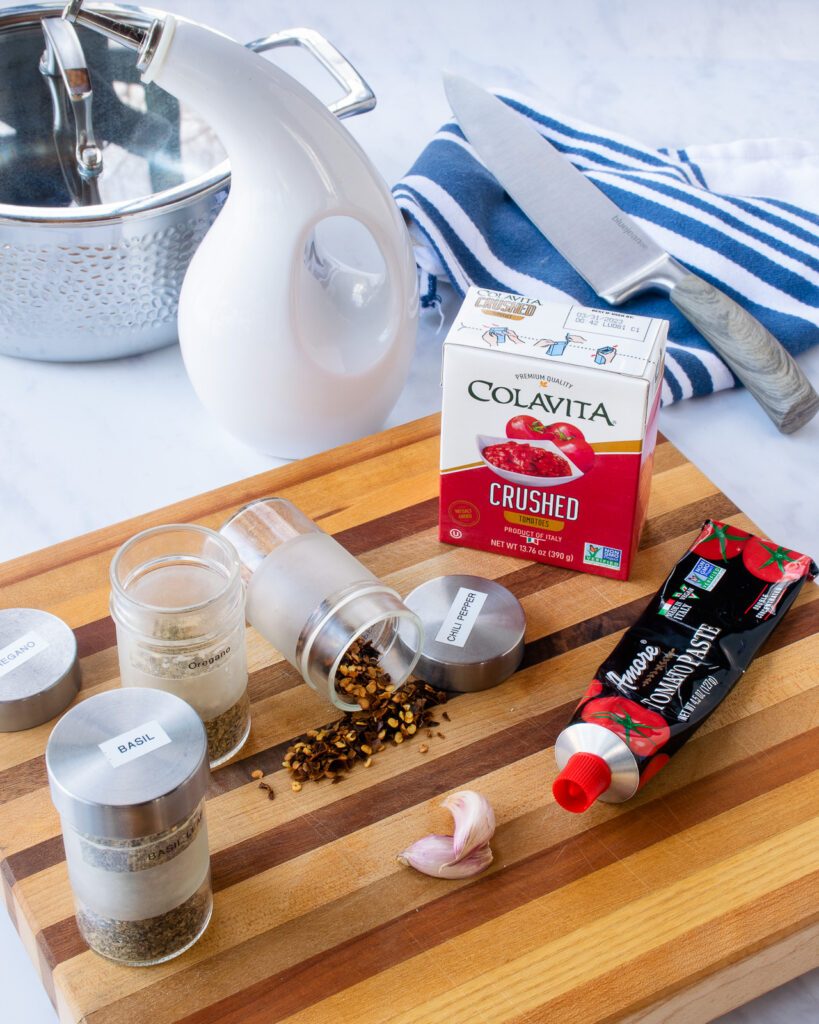 Ingredients on a cutting board - crushed tomatoes, tomato paste, chili flakes, basil, oregano and a saucepan, cruet of oil and chef's knife in the background.