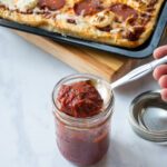 A spoon lifting pizza sauce out of a jar with a cooked pizza in the background.