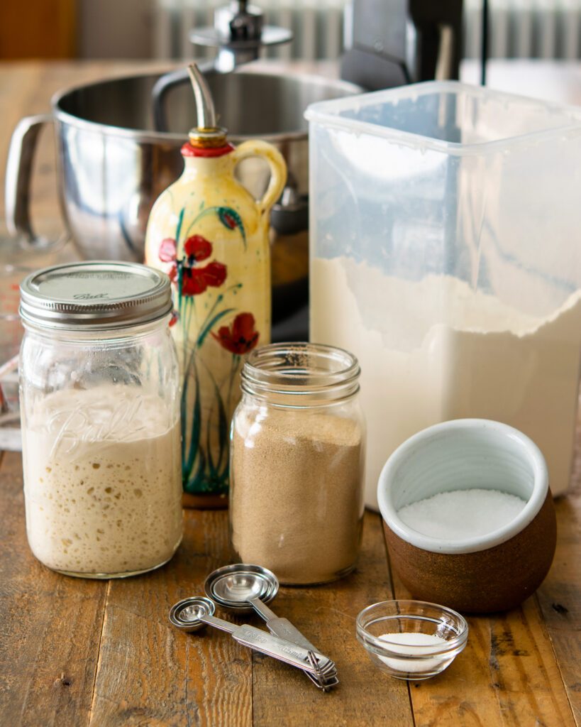 Ingredients on a table with a stand mixer in the background - sourdough starter, yeast, salt, oil, flour, sugar.