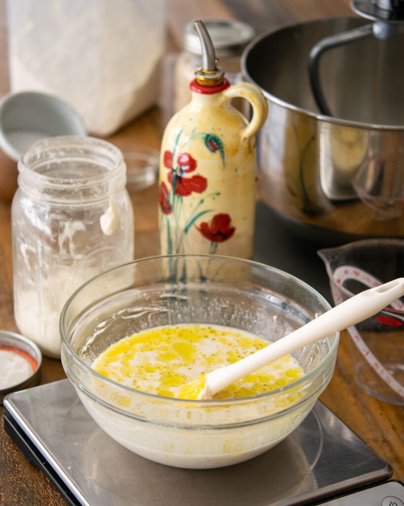 A bowl of liquid on a scale with other ingredients and a stand mixer in the background.