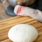 A ball of pizza dough on a cutting board with a stand mixer in the background.