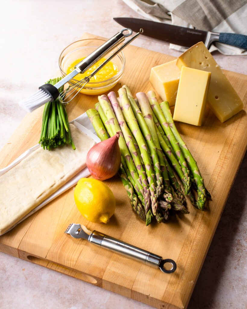 Ingredients on a cutting board - asparagus, lemon, shallot, three cheeses, chives, puff pastry and some beaten egg.