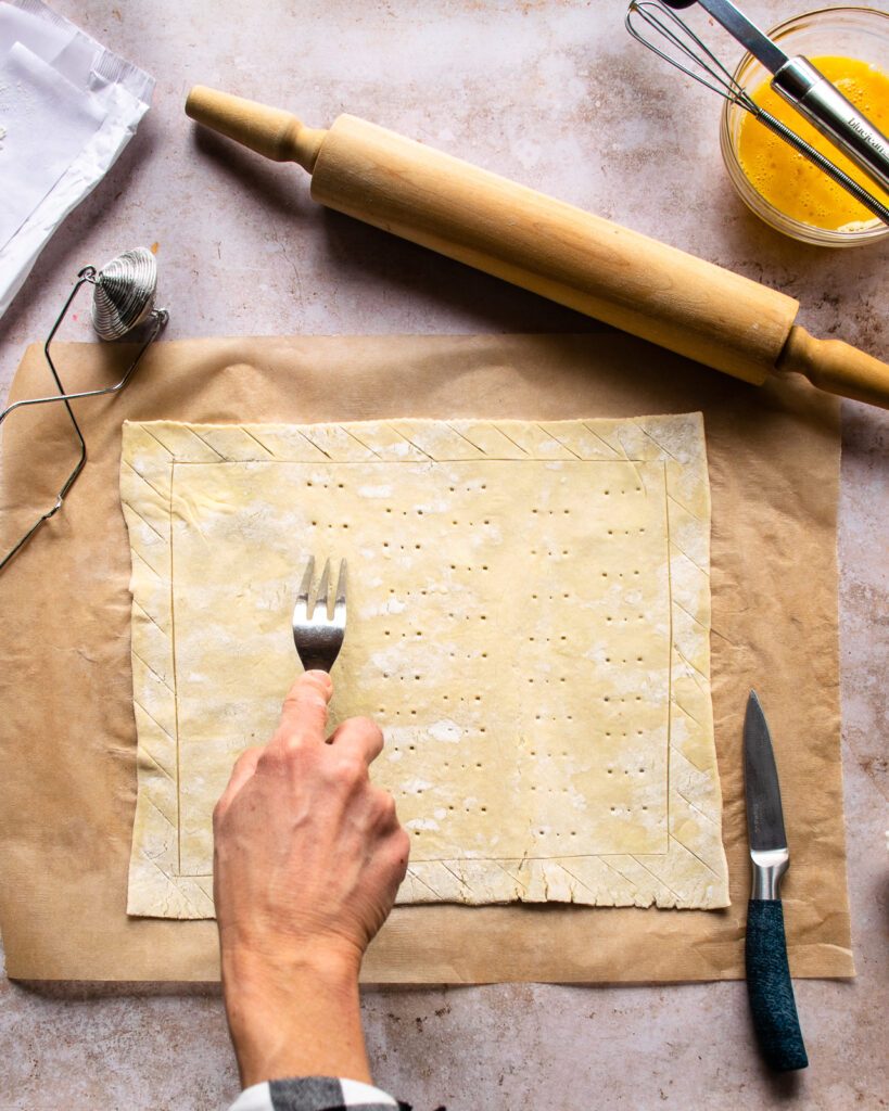 A hand pricking puff pastry with a fork, with a rolling pin and flour dusting wand nearby.