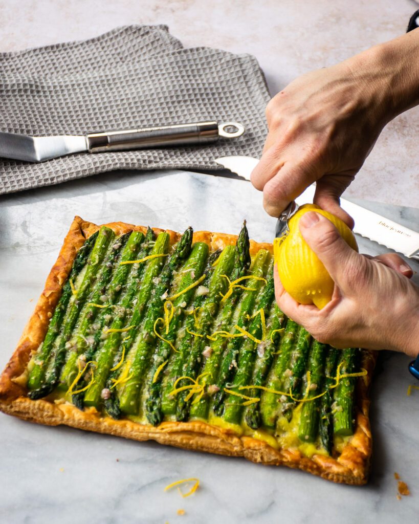 Hands zesting a lemon over an asparagus tart.