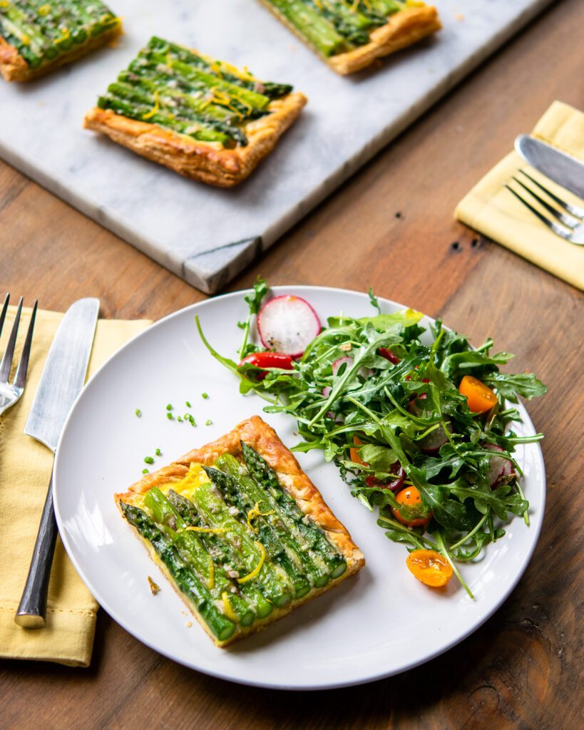 A piece of asparagus tart on a white plate with a side salad and more in the background.